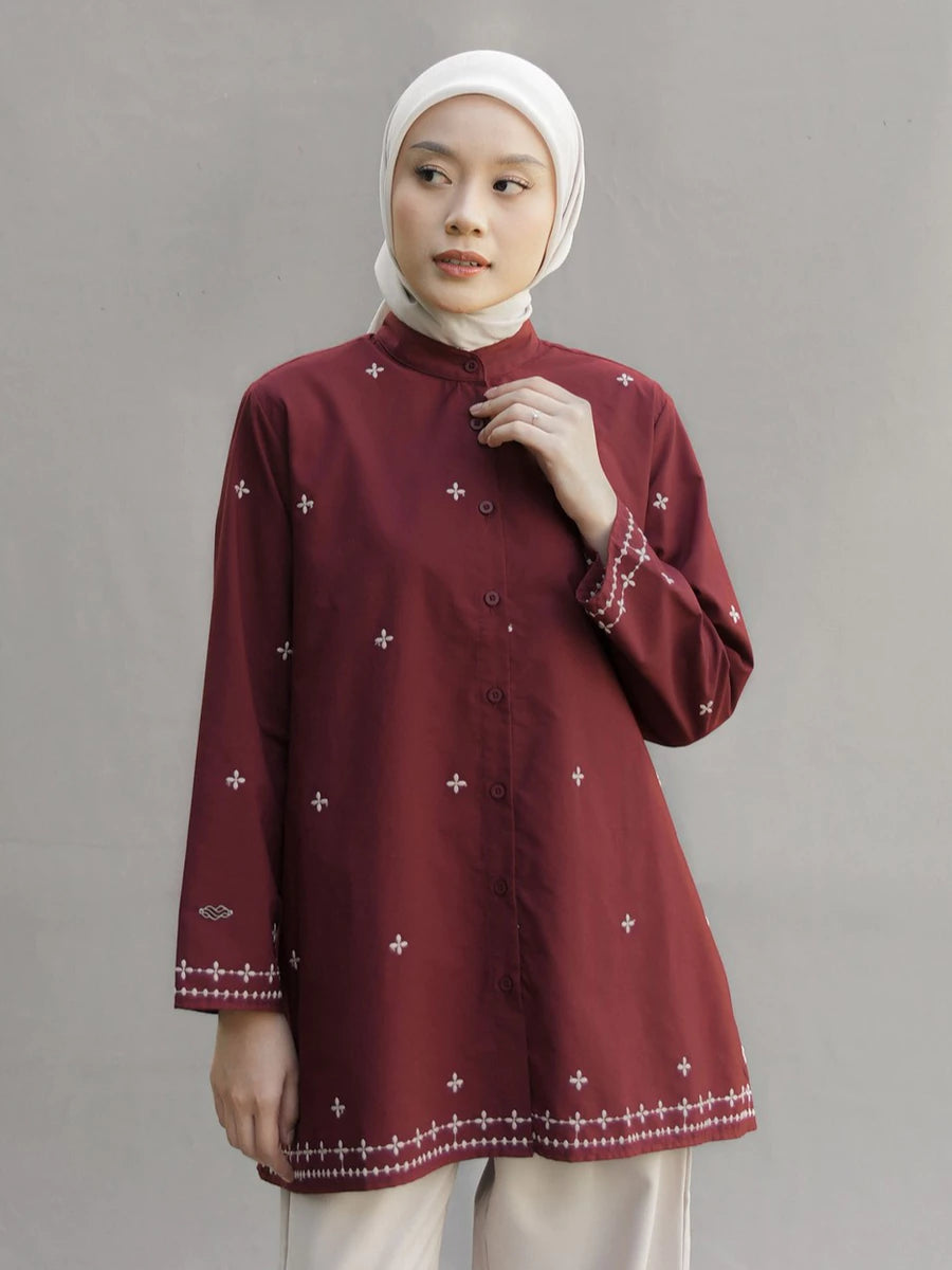 A woman wearing a maroon long-sleeve button-up tunic by Mistyvori with small white embroidered crosses and decorative borders, paired with a white hijab and light trousers, standing against a neutral gray studio background.