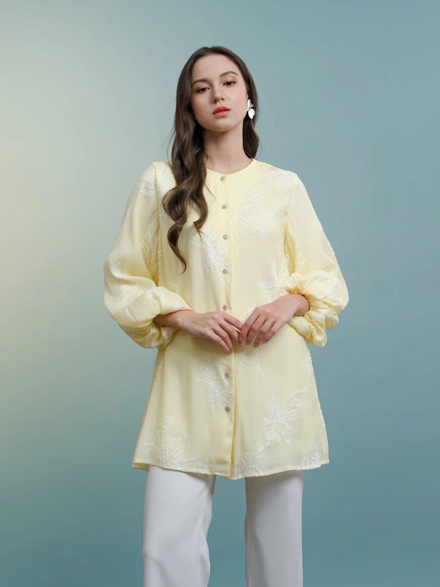 Woman wearing a light yellow blouse by Kasa Heritage with white pants against a gradient blue background