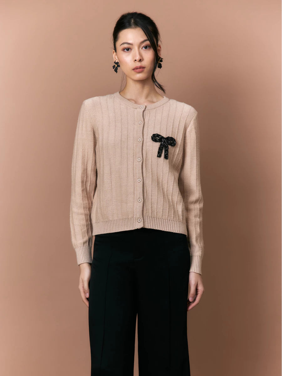 Woman wearing a beige cardigan by Studio Tui with a black bow detail on a brown background