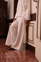 Person wearing a white top with lace details and linen pants by Kasyalia on a wooden floor.