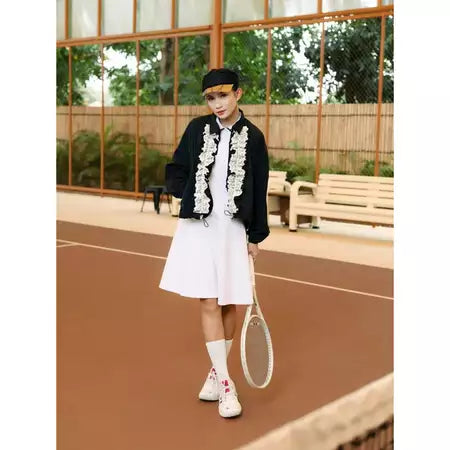 Person on a tennis court wearing a black jacket by Haidee Orlin with white floral embroidery and a white skirt.