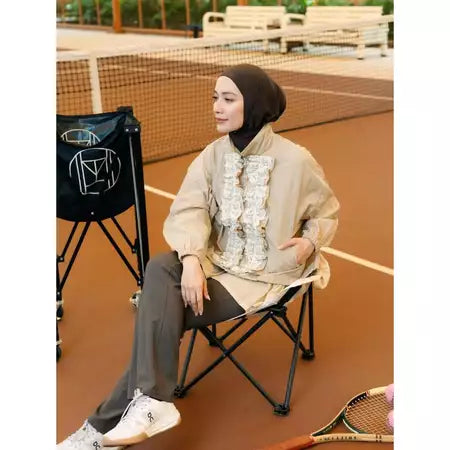 Person sitting on a chair on a tennis court wearing a beige jacket by Haidee Orlin with ruffled details.