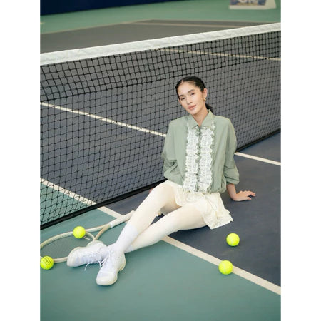 Woman sitting on a tennis court with tennis balls around her, wearing a sage green jacket by Haidee Orlin and white skirt.