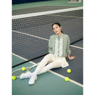 Woman sitting on a tennis court with tennis balls around her, wearing a sage green jacket by Haidee Orlin and white skirt.