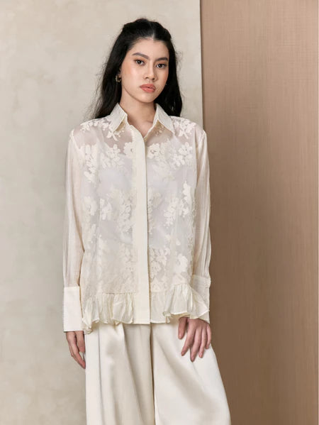 Woman wearing a white lace shirts by Studio Tui and white pants against a beige background