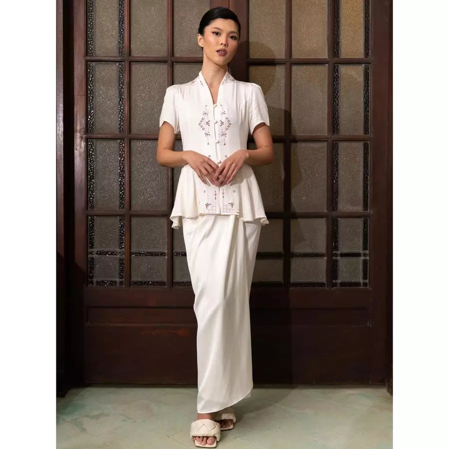 Woman wearing a traditional white kebaya by Kartinis Label standing against a wooden paneled wall.