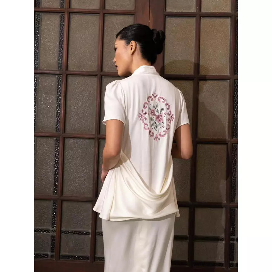 Woman wearing a white traditional kebaya by Kartinis Label with floral embroidery on the back, standing against a textured wall.