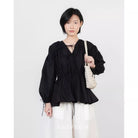 A model wearing a black long-sleeve blouse by Kasyalia with a V-neckline, tie details at the collar and cuffs, and a gathered peplum waist, paired with white drawstring pants and holding a cream woven shoulder bag.