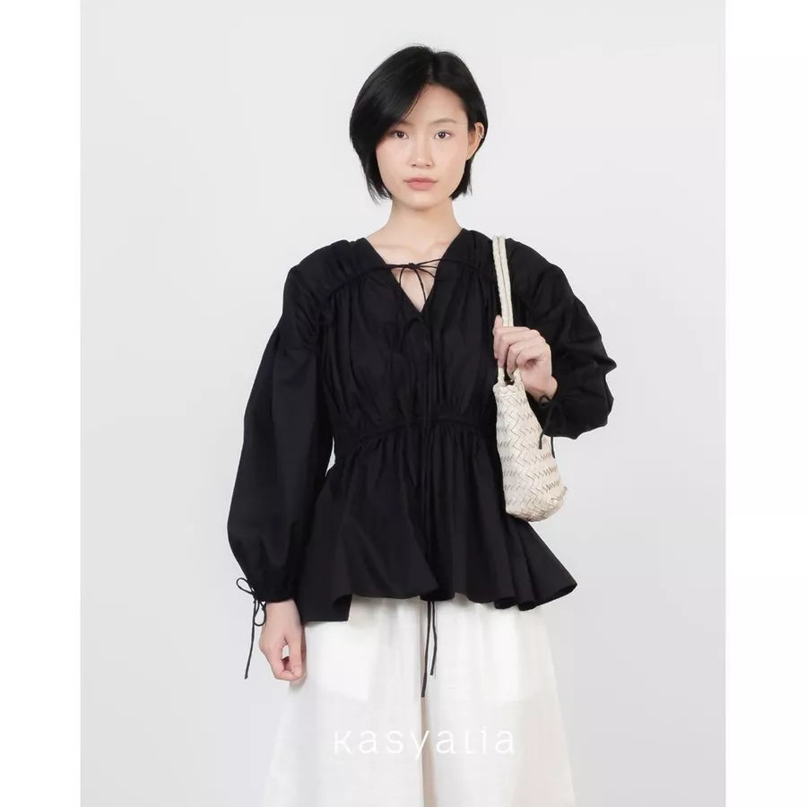 A model wearing a black long-sleeve blouse by Kasyalia with a V-neckline, tie details at the collar and cuffs, and a gathered peplum waist, paired with white drawstring pants and holding a cream woven shoulder bag.