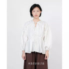 A model wearing a white long-sleeve blouse with a tie-front keyhole and gathered peplum waist, paired with dark brown wide-leg pants, against a plain light gray background. The brand name 'kasyalia' is printed at the bottom.