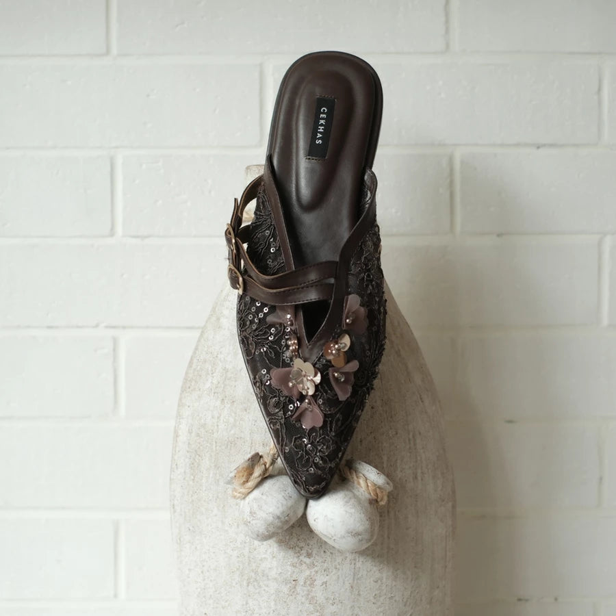 Brown mules sandal by Cekhas with floral embellishments on a textured surface with a white tiled background