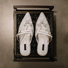 Pair of white pointed-toe mules sandals by Cekhas with floral designs in a decorative wooden box.