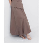 A woman wearing a brown Sienna skirt with soft pleats by Kasyalia, paired with a matching top. The skirt reaches down to the ankles and has a flowing silhouette.