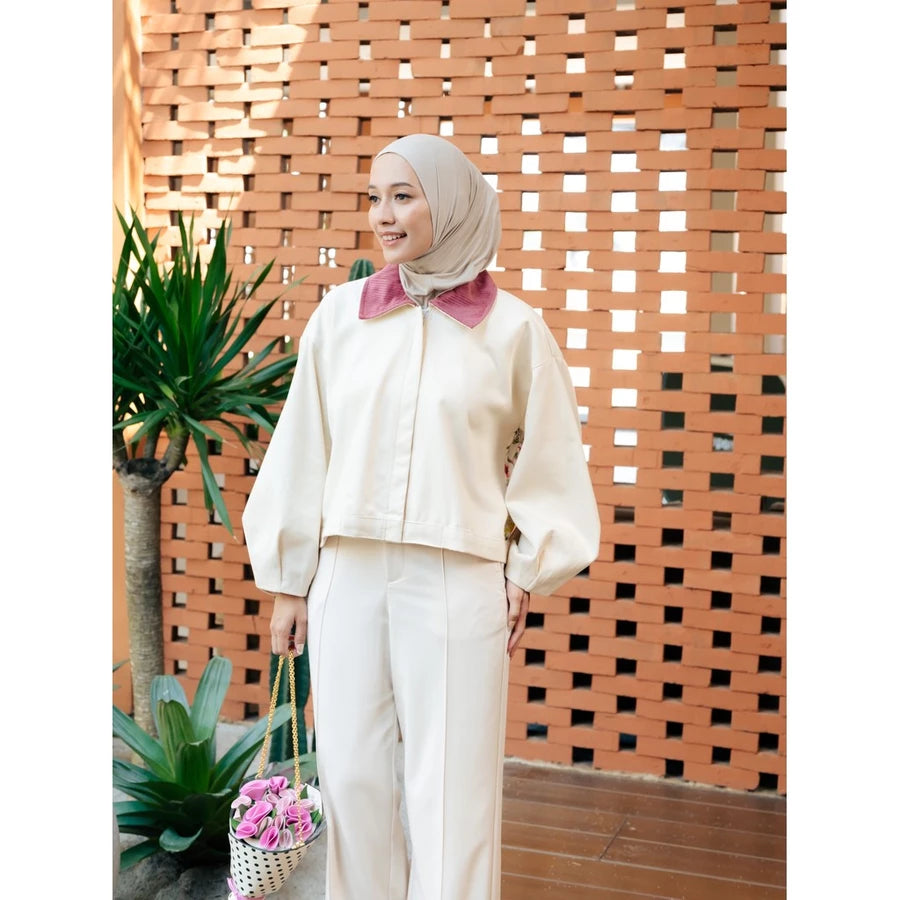 A woman wearing a cream-colored jacket by Haidee Orlin with a contrasting dusty pink collar, a beige hijab, and holding a small basket of pink flowers, standing in front of a terracotta lattice wall.