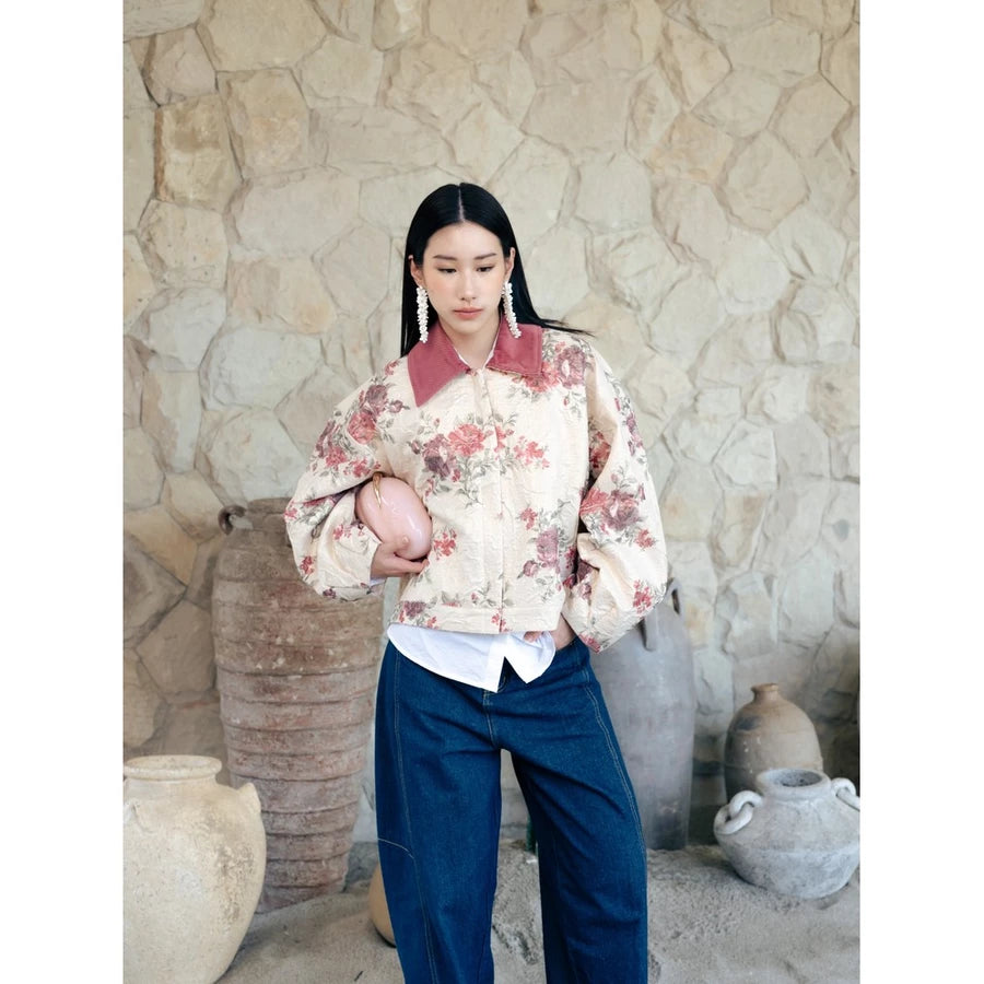 A woman stands in front of a rustic stone wall wearing a cream floral bomber jacket by Haidee Orlin with a dusty rose collar, dark blue wide-leg pants, and holding a pink clutch.