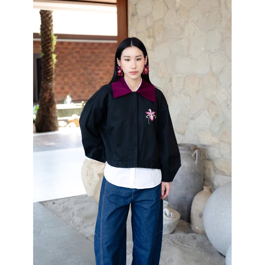 A woman stands in a stone-walled outdoor setting wearing a black cropped jacket by Haidee Orlin with a deep burgundy collar, a white shirt underneath, blue jeans, and large pink earrings.