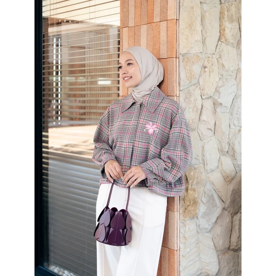 A woman wearing a light beige hijab and a pink and gray plaid long-sleeve jacket by Haidee Orlin with a pink bow accent, paired with white pants and holding a dark purple handbag, standing next to a stone wall.