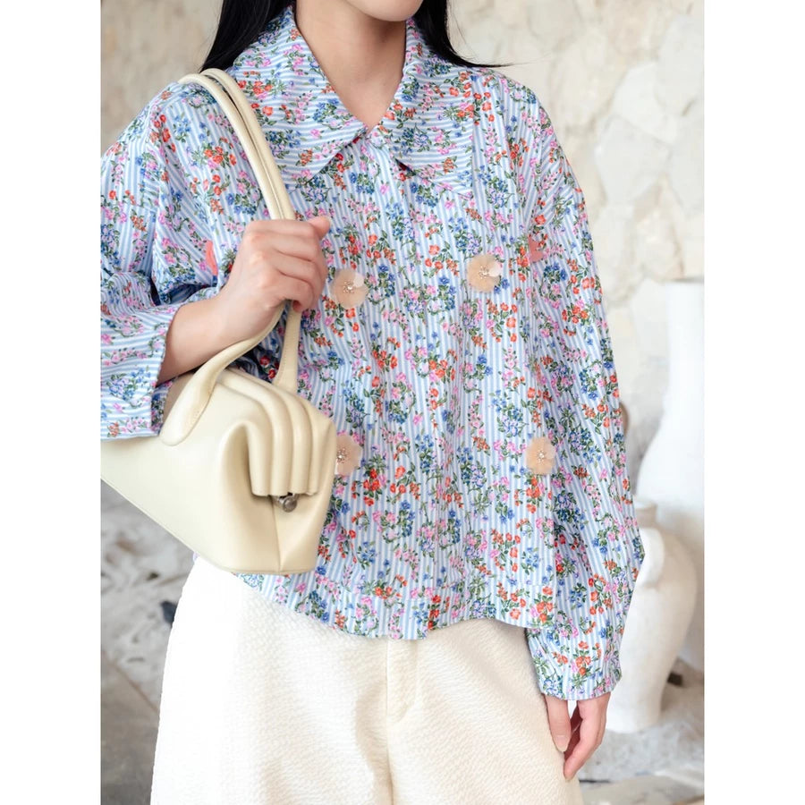 A person wearing a light blue and white floral long-sleeve jacket by Haidee Orlin with beige buttons decoration, paired with cream-colored pants and carrying a cream-colored shoulder bag.