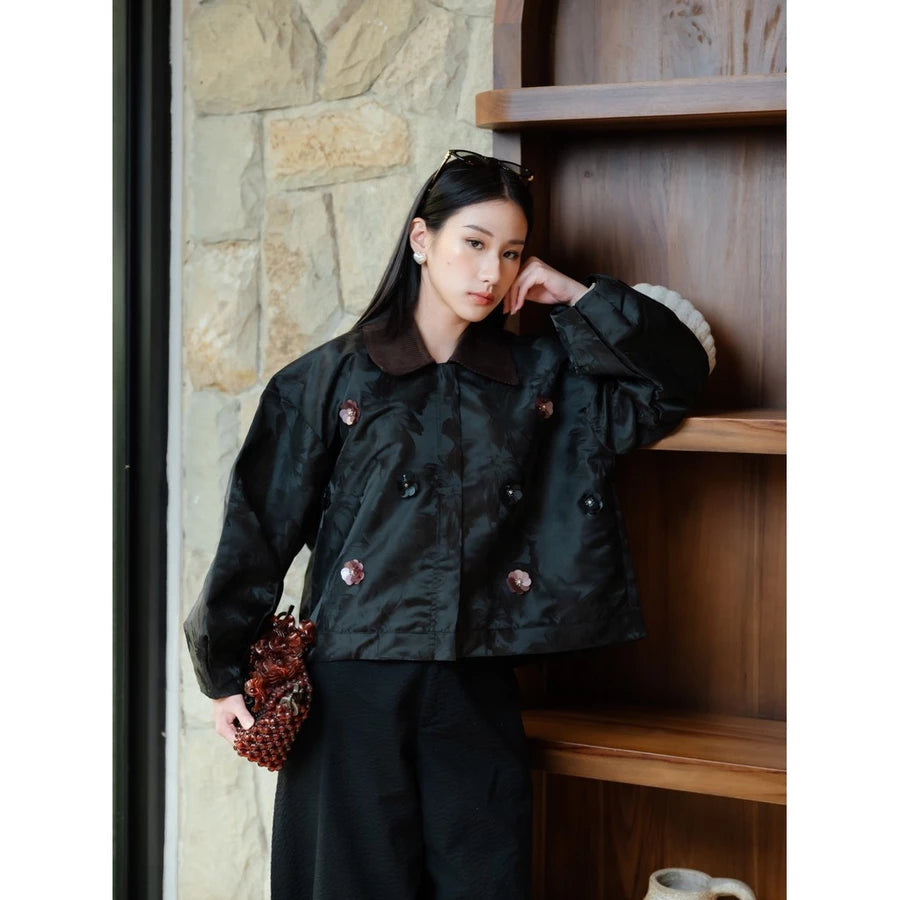 A woman models a black double-breasted jacket by Haidee Orlin with a dark brown collar and decorative buttons, paired with black trousers, standing next to a wooden shelf.