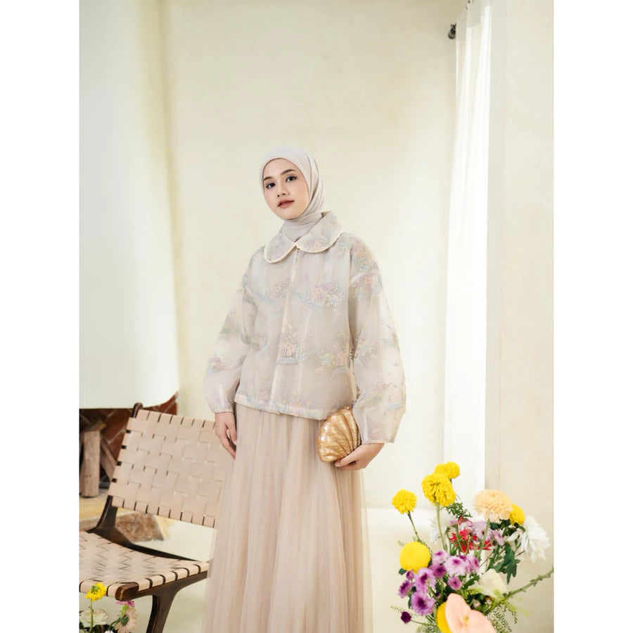 Woman wearing a pink lace outer with floral details by Haidee Orlin with a hijab standing indoors next to a chair and flowers.