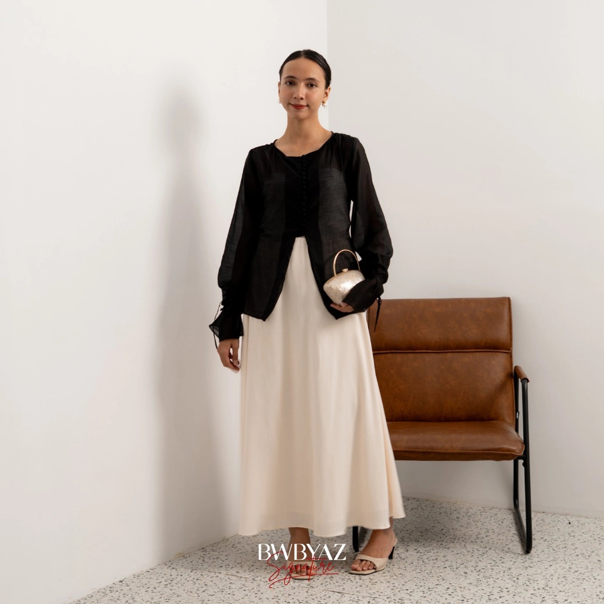 A woman wearing a black button top blouse from BWBYAZ and a sheer beige skirt, standing in a room with a chair and a wall in the background.