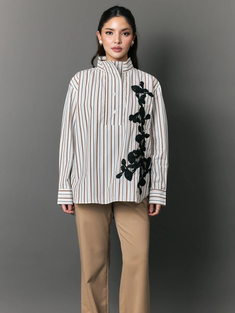 Woman wearing a striped shirt with black floral embroidery by Studio Tui and beige pants against a gray background