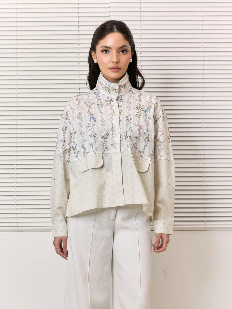 A woman stands in front of white window blinds wearing a high-neck, long-sleeve cropped shirt by Studio Tui with a floral embroidered pattern on the upper portion and solid light beige sleeves, paired with white trousers.