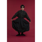 A model wearing a black long-sleeve blouse with white polka dots and a voluminous black skirt by Yestoday, posing against a solid dark red studio background.
