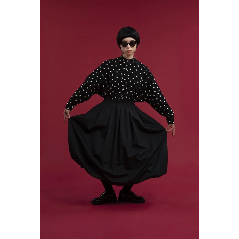 A model wearing a black long-sleeve blouse with white polka dots and a voluminous black skirt by Yestoday, posing against a solid dark red studio background.