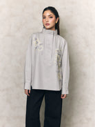 Woman wearing a light gray shirt with floral embroidery by Studio Tui and black pants against a beige background
