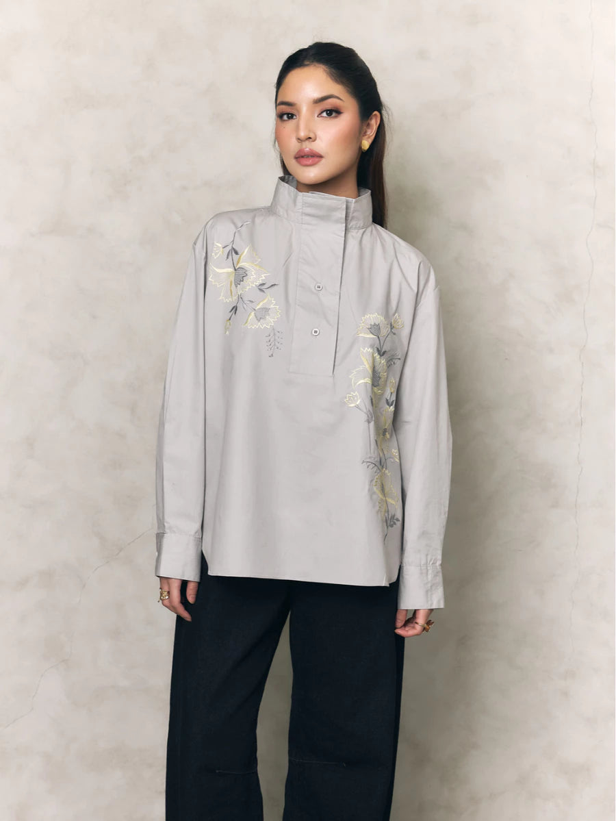 Woman wearing a light gray shirt with floral embroidery by Studio Tui and black pants against a beige background