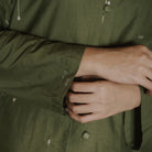 Close-up of a person wearing an olive green button-up tunic, showing the fabric texture, cuff detail with small white embroidered dots, and hands adjusting the shirt.