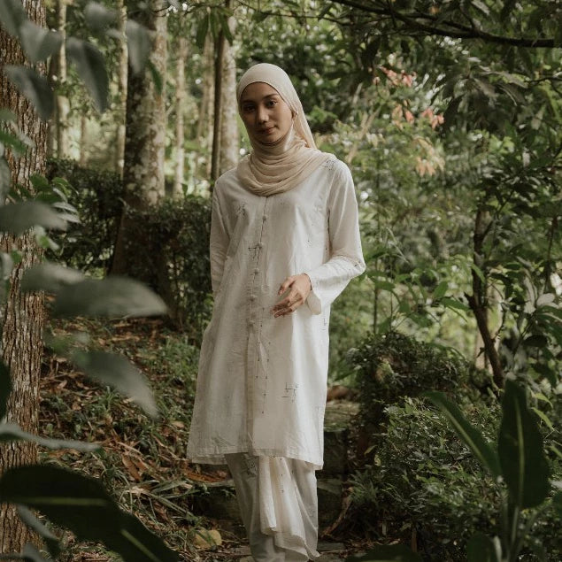 A woman wearing a white long-sleeve tunic and matching pants with a beige hijab stands in a lush green forest.