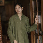 A woman wearing an olive green long-sleeve button-up tunic with white embroidered details, standing in a rustic indoor setting next to a wooden cabinet.