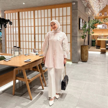 A woman wearing a pink tulip shirt with long sleeves by Casa Elana, paired with white trousers, standing in a room with wooden dividers and a table with chairs in the background.