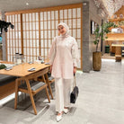 A woman wearing a pink tulip shirt with long sleeves by Casa Elana, paired with white trousers, standing in a room with wooden dividers and a table with chairs in the background.