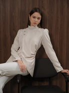 Woman wearing a light-colored blouse and white pants by Kasa Heritage sitting on a chair against a wooden wall.
