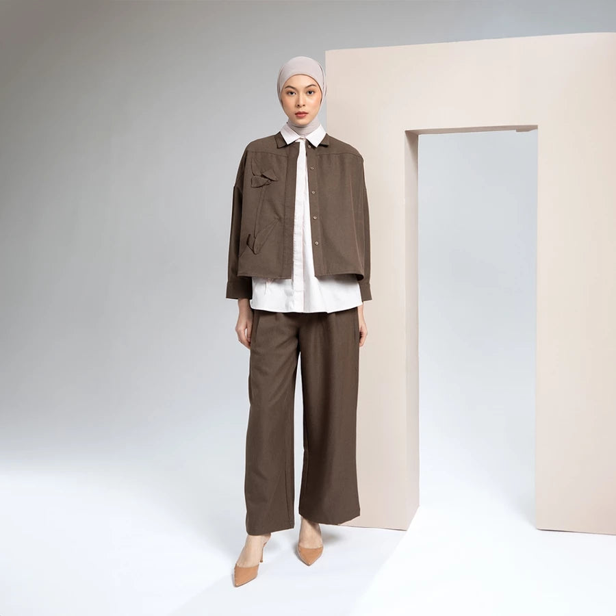 Person wearing a brown outer jacket by Hijabchic with a white shirt and beige hijab against a plain background