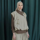 Woman wearing a beige vest by Jenna & Kaia and brown blouse against a dark green curtain background