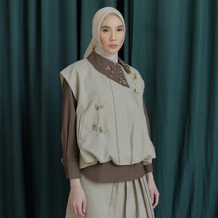 Woman wearing a beige vest by Jenna & Kaia and brown blouse against a dark green curtain background