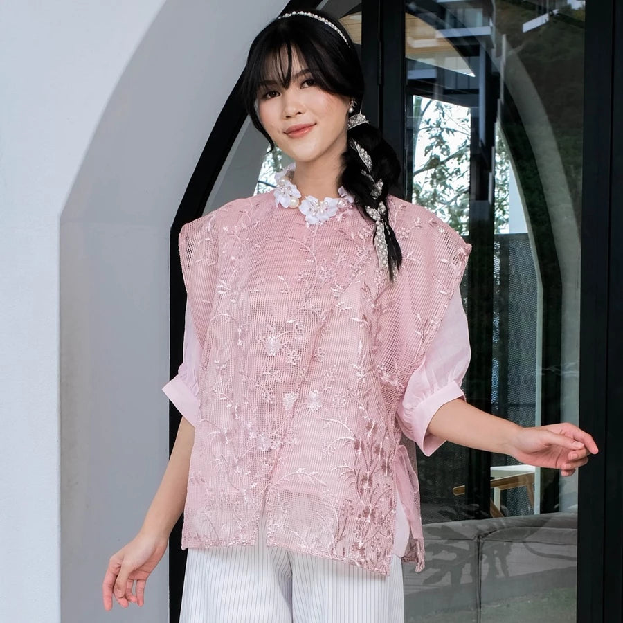 Woman wearing a pink embroidered lace outer by Holy Peony and white pants standing indoors.