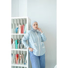 Woman wearing a light blue shirt by Abame with a hijab in front of a bookshelf.