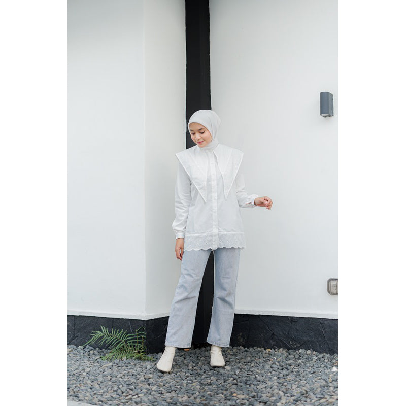 Woman wearing a white layered long-sleeve shirt by Casa Elana, light blue jeans, and a white hijab standing against a minimalist white wall.