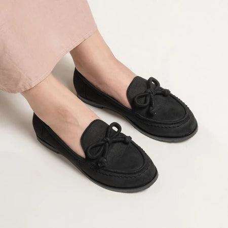 Black loafers flat shoes with bow details from Dianable worn by a person on a light background