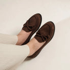 Brown loafers flat shoes with bow details from Dianable worn by a person in a beige sweater on a light background
