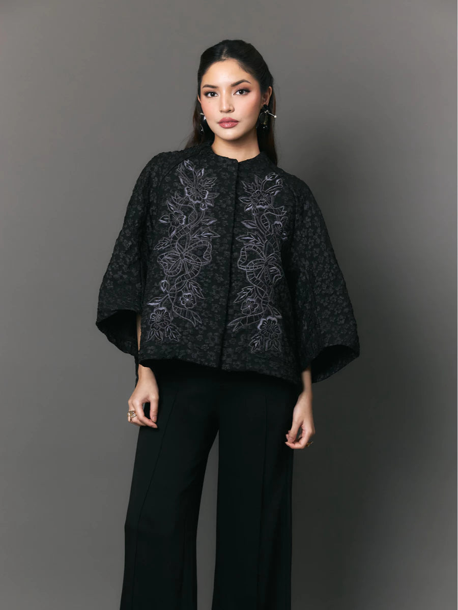 Woman wearing a black patterned outer with floral embriodery by Studio Tui against a gray background