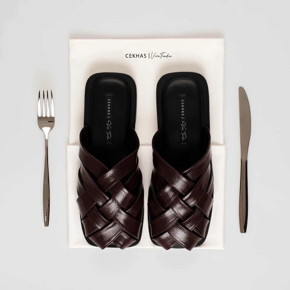 Pair of brown woven sandals by Cekhas in a box with a fork and knife on a light gray background
