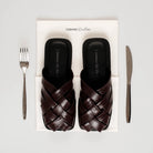 Pair of brown woven sandals by Cekhas in a box with a fork and knife on a light gray background