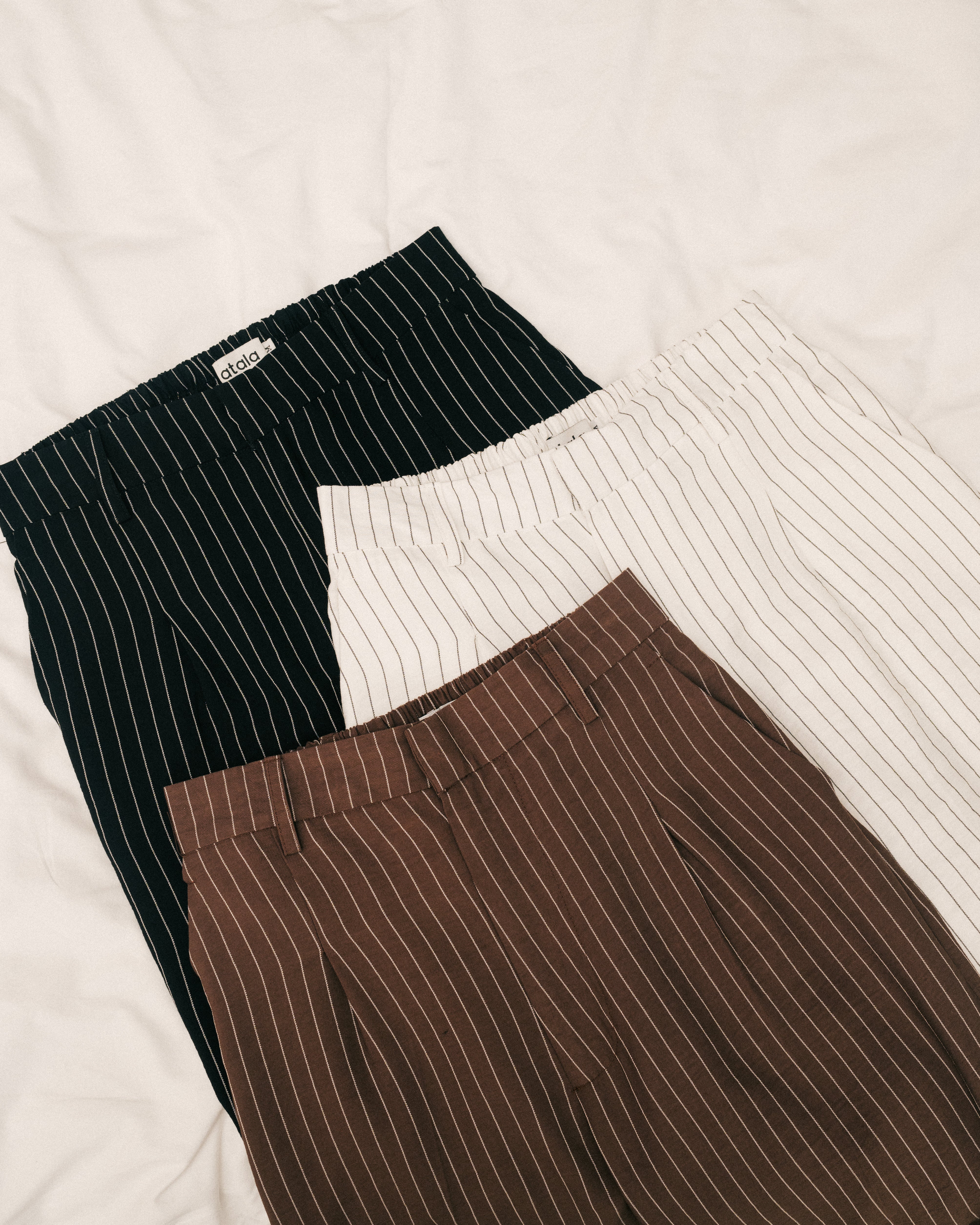 Three pairs of pinstripe pants in black, white, and brown from Atala Scarves on a light background.