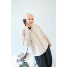 Woman wearing a beige hijab and shirt by Abame, holding a black phone receiver against a white background.